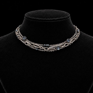 May include: A multi-strand necklace featuring a combination of silver-toned oval links and a box chain. The design is accented with small, dark blue circular crystals set in metal bezels, distributed evenly along the layered chain structure.