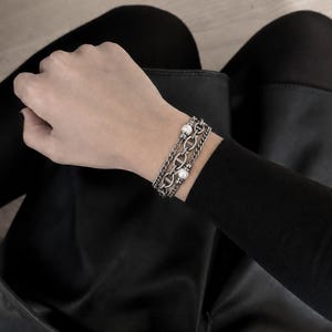 Könnte beinhalten: Ein mehrsträngiges silberfarbenes Armband mit einer zentralen Kette aus ovalen Gliedern, flankiert von zwei dünneren Panzerketten. Das Design ist mit zwei kleinen, runden weißen Perlen akzentuiert, die symmetrisch angeordnet sind.