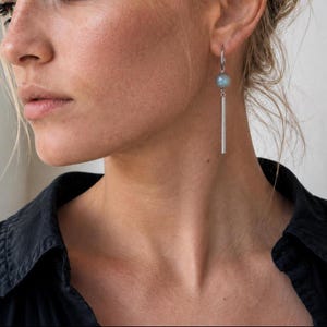 Peut inclure: Une boucle d'oreille pendante argentée composée d'un petit anneau circulaire en haut, d'une perle ronde en pierre gris-bleu suspendue en dessous, et d'une fine tige métallique argentée polie qui pend verticalement sous la perle.