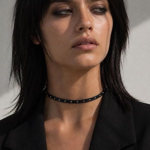 Może przedstawiać: Cienki czarny naszyjnik typu choker z rzędem małych, równomiernie rozmieszczonych srebrnych metalowych ćwieków na całej długości paska. Dodatek ściśle przylega do szyi, zapewniając prosty i jednolity wygląd całego elementu biżuterii.