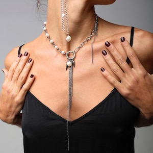 May include: A silver-toned necklace with a pearl and chain design. The necklace features a large chain link section, a circular pendant, and a long chain tassel. A matching earring with a pearl and chain is also visible.