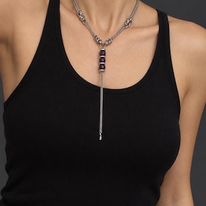 Könnte beinhalten: Eine silberfarbene Lariat-Halskette mit geflochtenem Kettendesign und drei dunkelvioletten facettierten Perlen, die vertikal in der Mitte angeordnet sind, abgeschlossen mit einem dünnen hängenden Silberkettchen am unteren Ende.
