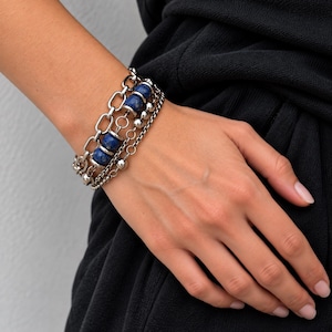 May include: A multi-strand bracelet featuring a combination of silver-toned metal chains and smooth, round, deep blue lapis lazuli beads. The design incorporates a mix of thick oval links and thinner, delicate chains with small metallic spheres.