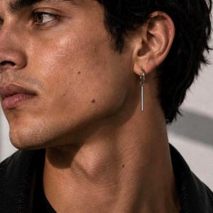 Op de afbeelding: Een zilverkleurige oorring met een dunne, verticale staafhanger. De oorbel wordt in de oorlel gedragen en heeft een gepolijste metalen afwerking met een eenvoudig, strak ontwerp dat recht naar beneden hangt vanaf de ronde oorring.