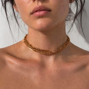 Puede incluir: Un collar tipo gargantilla de tono dorado con varias capas, que combina una cadena fina de cuentas con una cadena de eslabones ovalados alargados. El collar se ajusta al cuello y presenta un acabado metálico pulido con dos estilos de cadena.