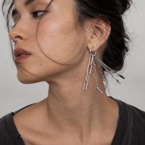 May include: A silver-toned hoop earring featuring multiple dangling attachments, including a long paperclip chain, a delicate link chain with small spherical beads, a thin chain with a flat circular charm, and a chain with dark rectangular beads.