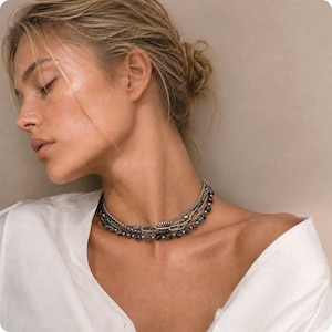 May include: A layered necklace featuring a combination of silver-toned metal chains and a strand of dark, faceted beads. The design includes a mix of delicate link chains and a thicker, elongated oval link chain, creating a textured, multi-strand look.