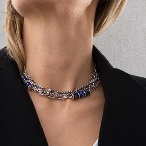 May include: A multi-strand silver-toned chain necklace featuring polished blue lapis lazuli beads interspersed with metallic spacers. The layered design consists of a delicate cable chain and a thicker link chain, resting around the neck area.