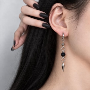 May include: A silver-toned dangle earring featuring a circular hoop, a small metallic sphere, a larger black spherical bead, and a pointed conical charm at the base. The earring is constructed with interconnected silver-coloured metal rings.