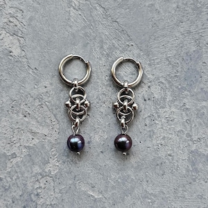 Op de afbeelding: Een paar zilverkleurige oorringen met een centraal decoratief element bestaande uit drie in elkaar grijpende metalen ringen. Elke oorbel is afgewerkt met een enkele, ronde, donkerpaars-zwarte parel die onderaan de metalen structuur hangt.