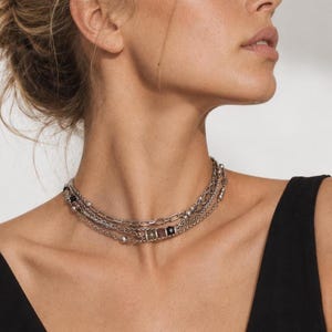 Peut inclure: Un collier multi-rangs de couleur argentée composé de fines chaînes à maillons, de petites perles métalliques et d'un élément central formé de trois petites perles rectangulaires aux tons terreux et noir, porté autour du cou.