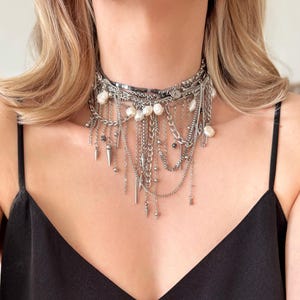 May include: A layered silver-tone metal choker necklace featuring a mix of delicate chains, small metallic beads, and irregular white freshwater pearls. The design includes multiple dangling elements, including thin metal spikes and various chain lengths.