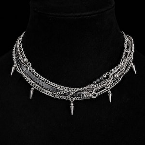Puede incluir: Un collar de cadenas m&uacute;ltiples en tono plateado que presenta una combinaci&oacute;n de cadenas de eslabones finos, una fila de cuentas met&aacute;licas cuadradas y varios pinchos c&oacute;nicos colgantes. El collar est&aacute; construido con diversas texturas de cadena.