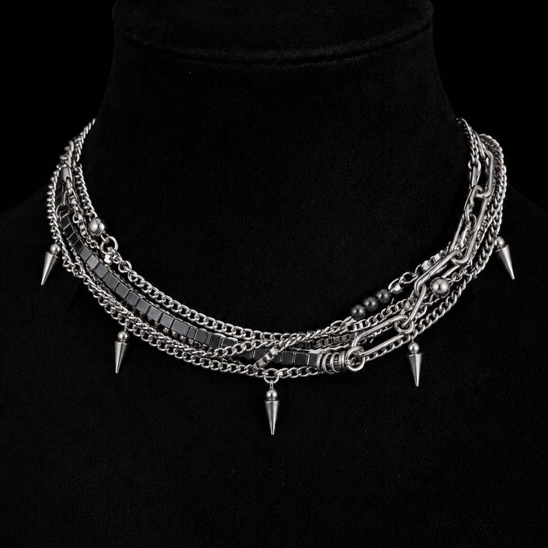Puede incluir: Un collar de cadenas m&uacute;ltiples en tono plateado que presenta una combinaci&oacute;n de cadenas de eslabones finos, una fila de cuentas met&aacute;licas cuadradas y varios pinchos c&oacute;nicos colgantes. El collar est&aacute; construido con diversas texturas de cadena.