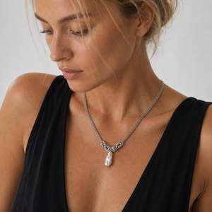 May include: A silver-toned chain necklace featuring a woven metal link design and a single, irregular white baroque pearl pendant suspended at the center. The necklace rests against the collarbone, showcasing a metallic texture and a lustrous pearl.