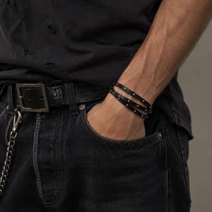 Pode incluir: Pulseira de couro preta com tachas prateadas. A pulseira está enrolada no pulso. A imagem também mostra um cinto preto, jeans pretos e uma camiseta preta.