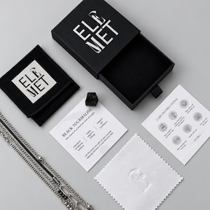 Puede incluir: Un joyero negro con el logotipo de ELEMET, una bolsa negra, un collar de cadena plateada, una piedra de turmalina negra en bruto, una tarjeta informativa sobre la turmalina negra, una tarjeta de instrucciones de cuidado y un pa&ntilde;o blanco.