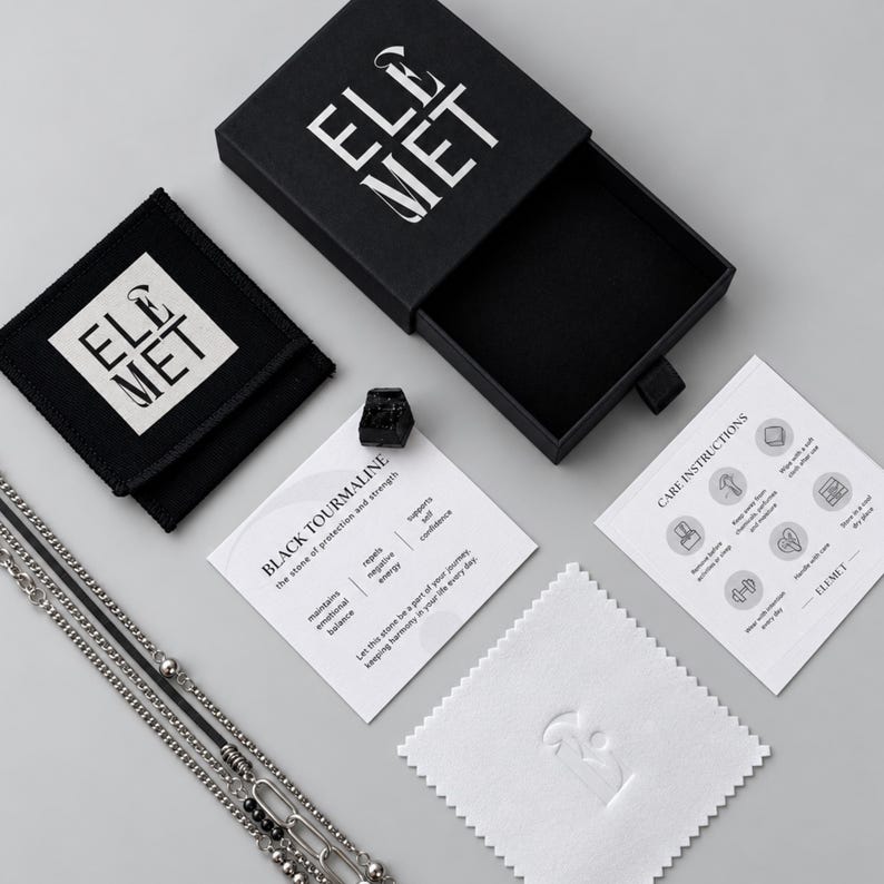 Puede incluir: Un joyero negro con el logotipo de ELEMET, una bolsa negra, un collar de cadena plateada, una piedra de turmalina negra en bruto, una tarjeta informativa sobre la turmalina negra, una tarjeta de instrucciones de cuidado y un pa&ntilde;o blanco.