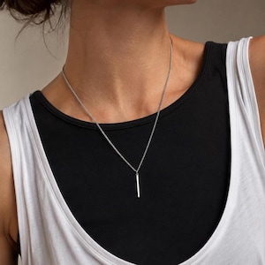Puede incluir: Un collar de tono plateado que presenta una cadena delicada y un colgante de barra rectangular vertical y delgada. La barra de metal cuelga centrada de la cadena, descansando sobre una prenda superior superpuesta en blanco y negro.