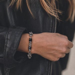 Könnte beinhalten: Ein Armband mit einer Reihe von polierten, silberfarbenen Metallgliedern, durchsetzt mit glatten, runden schwarzen Perlen und einer einzelnen durchscheinenden grauen Perle. Das Design enthält komplexe, strukturierte Metallteile an beiden Enden.