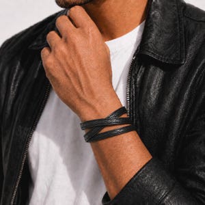 May include: A black leather wrap bracelet featuring a textured, cross-hatched pattern along the band. The bracelet is designed to wrap around the wrist multiple times, creating a layered appearance with a consistent matte black finish throughout.