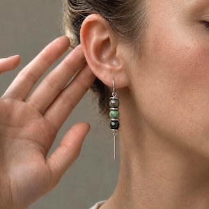 May include: A silver-toned drop earring featuring three stacked, faceted spherical beads in shades of mottled pink, green, and black, separated by small silver-toned spacers, and finished with a thin, straight silver-toned metal bar at the base.