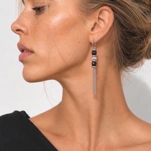 May include: A silver-toned dangle earring featuring three stacked beads, including two faceted black beads and one central marbled grey bead, finished with a delicate silver-toned chain tassel hanging from the bottom of the bead stack.