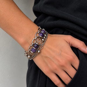 Pode incluir: Uma pulseira de várias camadas que combina correntes de metal prateado e contas roxas lisas e redondas. O design incorpora uma corrente de elos ovais grossos ao lado de uma corrente mais fina e delicada com pequenas esferas metálicas.