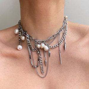 Puede incluir: Un collar de cadenas superpuestas de metal plateado con varios estilos de eslabones, incluyendo eslabones tipo barbada y caja. El collar está adornado con varias perlas de agua dulce blancas de forma irregular y pequeños pinchos metálicos.