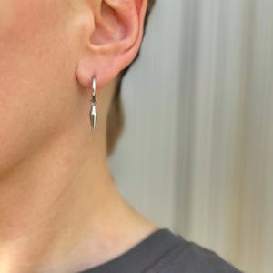 Puede incluir: Un pendiente de aro plateado con un colgante de punta plateada. El pendiente se lleva en el lóbulo de la oreja izquierda.