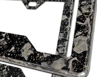 May include: A rectangular license plate frame featuring a black base with a marbled pattern of white and gray streaks, accented by small gold-colored flecks throughout the surface. The frame has a smooth, glossy finish with mounting holes.