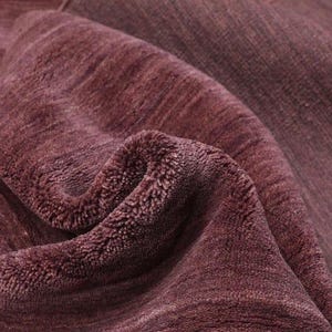 May include: Close-up of a folded, textured rug in a deep burgundy color. The rug's surface has a soft, plush appearance with visible fibers and a subtle striped pattern. The folds create shadows, enhancing the texture.