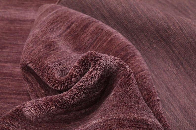 May include: Close-up of a folded, textured rug in a deep burgundy color. The rug's surface has a soft, plush appearance with visible fibers and a subtle striped pattern. The folds create shadows, enhancing the texture.