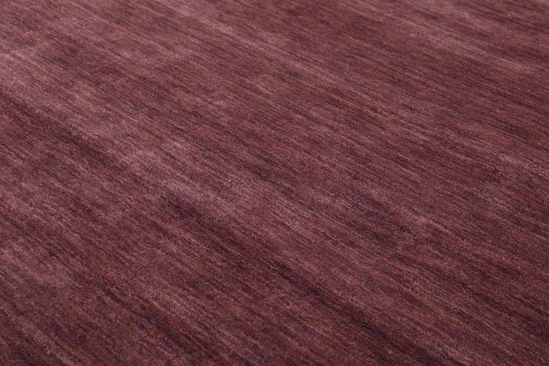 May include: Close-up of a burgundy-colored rug with a textured surface. The rug's fibers are arranged in a diagonal pattern, creating a sense of depth and movement. The color is a deep, rich shade of red-purple.