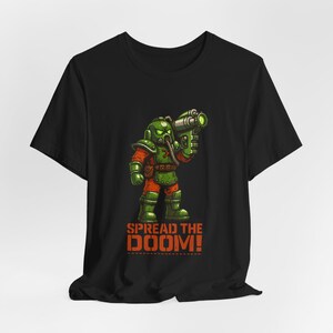 May include: Black t-shirt featuring a cartoon illustration of a character in a green gas mask and armor, holding a futuristic weapon. The text "SPREAD THE DOOM!" is printed in orange below the character. The design is a bold, eye-catching graphic.
