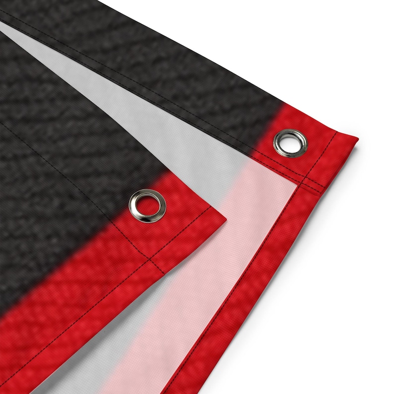 May include: Close-up of a flag with a black textured background, a white stripe, and a red border. The flag has metal grommets in the corners for hanging. The flag is made of a durable material.