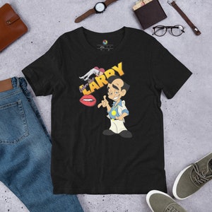 Puede incluir: Camiseta negra con una ilustración de dibujos animados de un hombre con el texto "Love that LARRY" encima. El hombre lleva una camisa blanca, una corbata amarilla y pantalones blancos. Sostiene un círculo amarillo en su mano derecha y tiene una expresión de sorpresa en su rostro.
