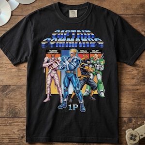 May include: A black short-sleeve t-shirt featuring a graphic print of the video game Captain Commando. The design displays the title text and four characters labeled Mummy Commando, Ninja Commando, and Baby Commando, with the text 1P below.