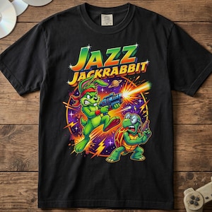 May include: A black short-sleeve t-shirt featuring a graphic print of the Jazz Jackrabbit logo and characters. The design shows a green rabbit and a turtle holding futuristic blasters against a purple and orange circular background.