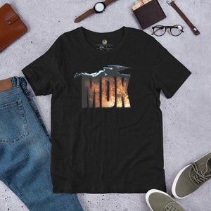 May include: Black short-sleeved t-shirt featuring the word "MDK" in a distressed, fiery font. The shirt is made of a soft material and is displayed on a flat surface with jeans, shoes, and accessories.