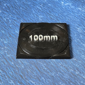 May include: A black square base with a round, black surface and the text "100mm" printed in white.