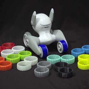 Może przedstawiać: Biały robot z niebieskimi kołami i różnymi kolorowymi kołami zamiennymi. Robot ma zaokrągloną głowę z dwoma antenami i szary korpus. Koła zamienne są w różnych kolorach, w tym czerwonym, zielonym, niebieskim, szarym i żółtym.