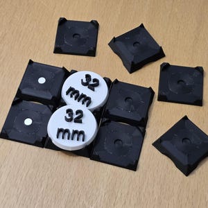 Puede incluir: Ocho cuadrados de plástico negro con un pequeño agujero en el centro, dispuestos en forma de cuadrícula. Dos círculos blancos con el texto "32 mm" se colocan encima de los cuadrados.
