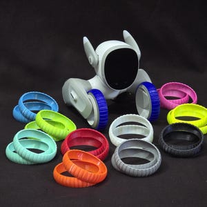 Könnte beinhalten: Ein weißes Roboter-Spielzeug mit blauen Rädern und einem schwarzen Bildschirm. Der Roboter ist umgeben von 10 bunten Silikonringen in verschiedenen Farben, darunter Rot, Orange, Gelb, Grün, Blau, Lila, Pink und Grau.