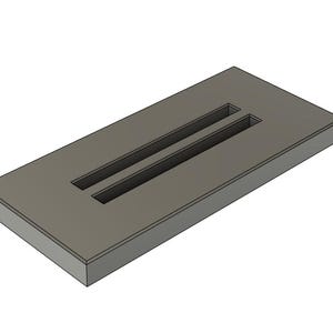 Könnte beinhalten: Ein rechteckiges, graues Objekt mit zwei parallelen, vertieften Schlitzen. Das Objekt hat eine abgeschrägte Kante, die ihm ein leicht abgewinkeltes Aussehen verleiht. Das Design ist einfach und geometrisch, mit klaren Linien und minimalistischer Ästhetik.