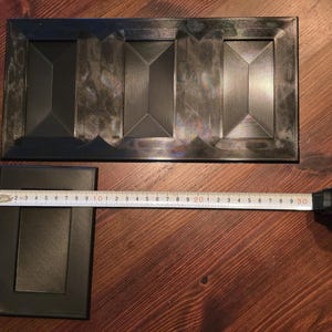 May include: Two dark gray rectangular decorative panels with geometric designs. A measuring tape shows the smaller panel is approximately 6 inches long. The larger panel has three geometric sections.