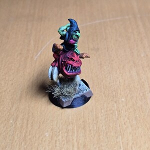 May include: A green goblin riding a red and black beast with a toothy grin. The goblin is wearing a black hooded cloak and is holding a weapon. The beast is standing on a brown base with green grass.