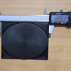 Könnte beinhalten: Ein schwarzes quadratisches Kunststoffteil mit einer runden, schwarzen, versenkten Scheibe in der Mitte. Eine digitale Schieblehre zeigt 12,00 mm an.