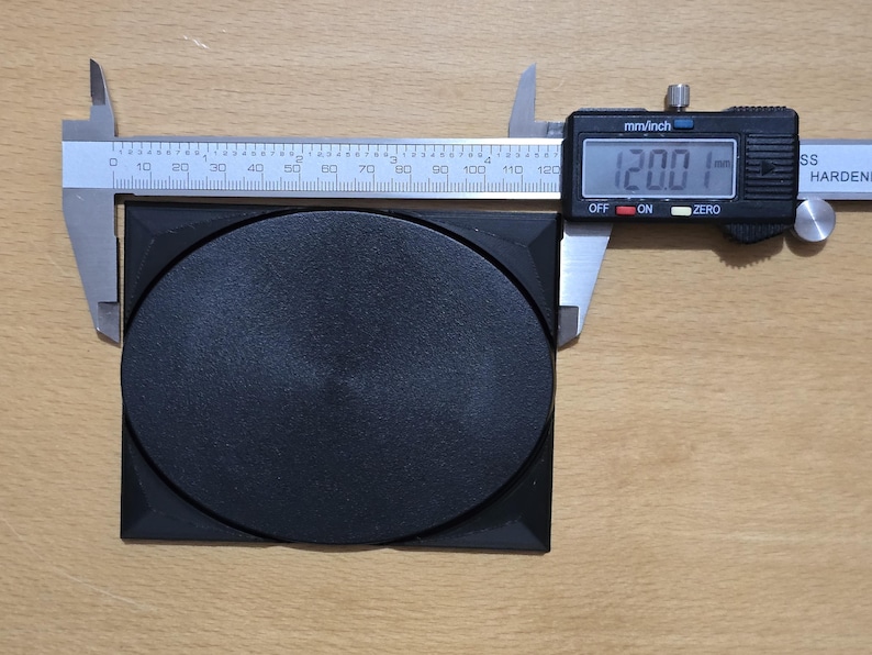 Op de afbeelding: Een zwarte vierkante kunststof met een ronde, zwarte, ingezonken schijf in het midden. Een digitale schuifmaat geeft 12,00 mm aan.