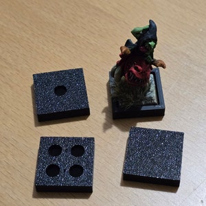 May include: Four black square bases with different numbers of holes. One base has one hole, another has three holes, and the other two are solid. A miniature figure of a goblin riding a beast is on a black base.